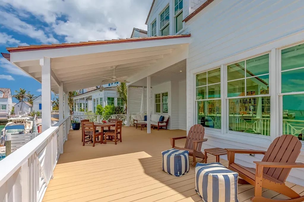 Deck with wooden floor, dining table, Adirondack chairs, and large windows overlooking palm trees.