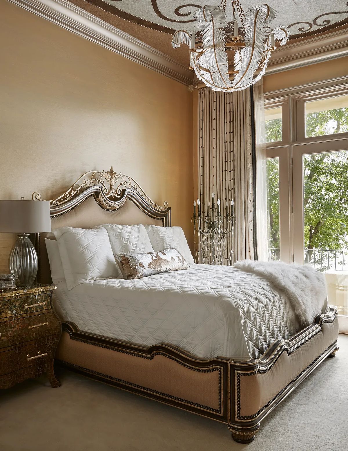 Bedroom with ornate bed, decorative nightstands, chandelier, gold walls, and tall windows with drapes.