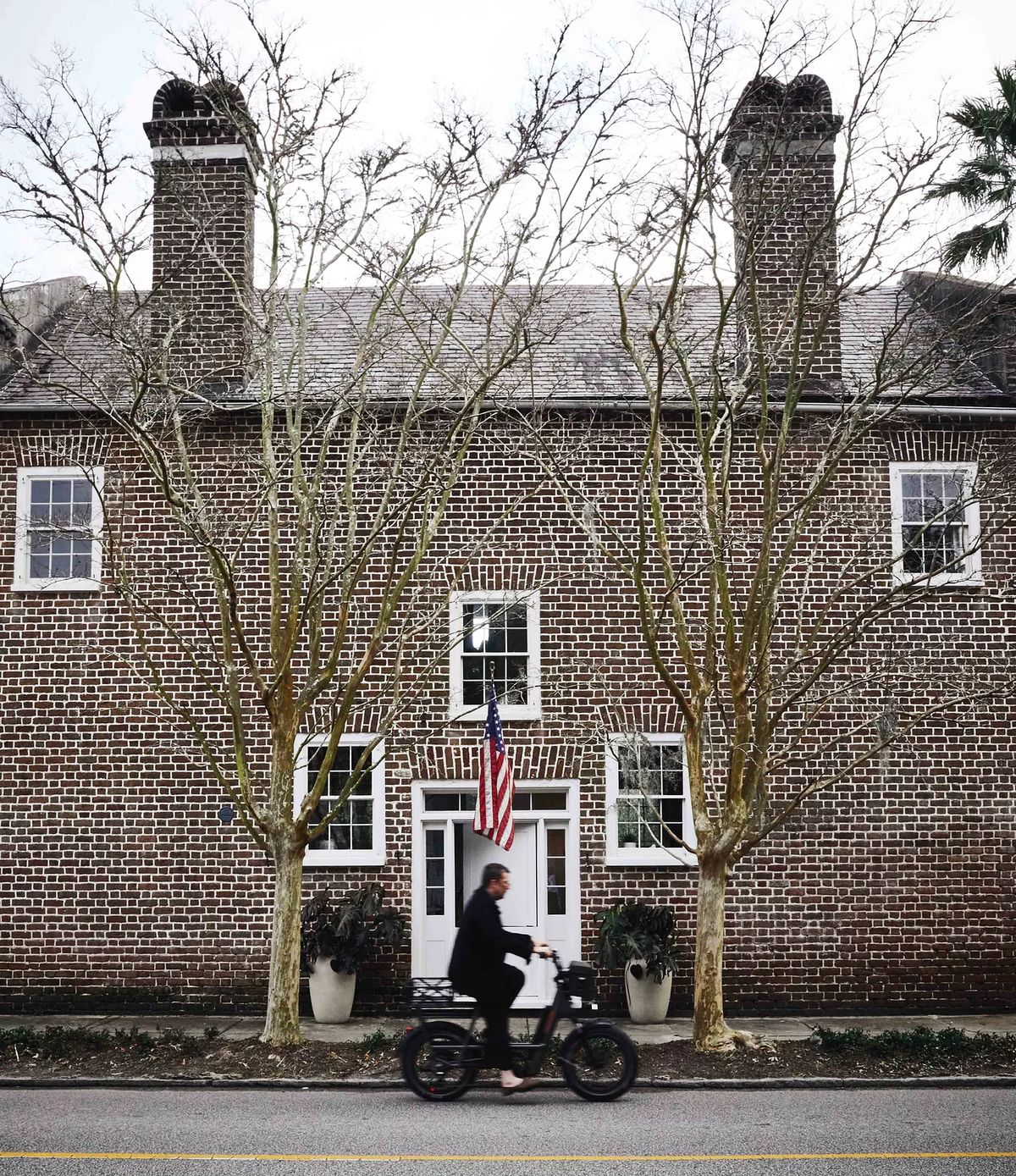 Front exterior of brick house with windows, trees, American flag, and person on motorcycle.