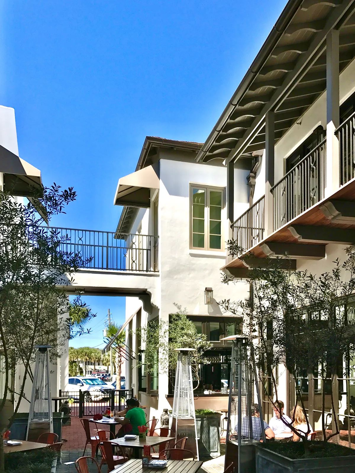 Outdoor patio with metal tables, chairs, stone flooring, greenery, and access to building and balcony above.