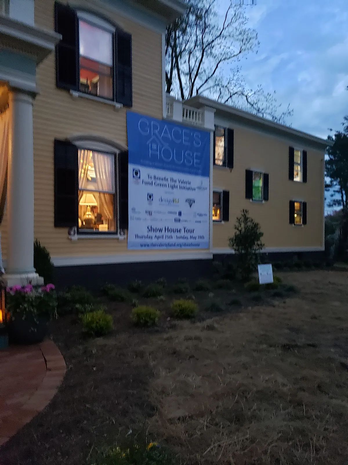 Exterior view of a yellow house with black shutters, illuminated windows, and a banner.
