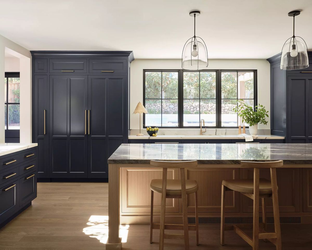 Kitchen with blue cabinetry, marble island, wooden bar stools, and large windows with pendant lights