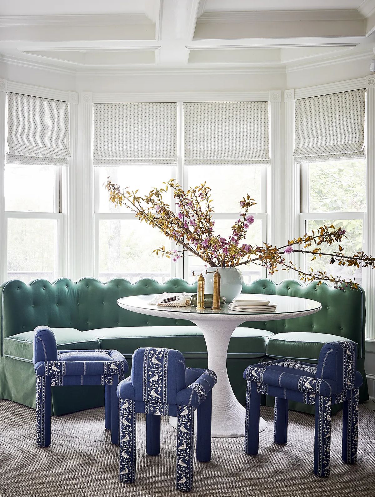 Dining room with round table, green banquette, blue chairs, and large vase with branches.