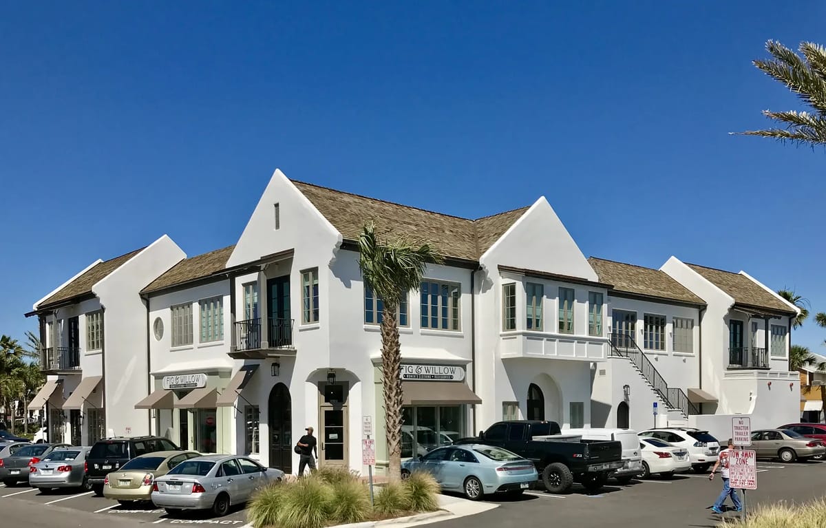 Commercial building with white exterior, brown sloped roof, multiple windows, awnings, and palm trees.