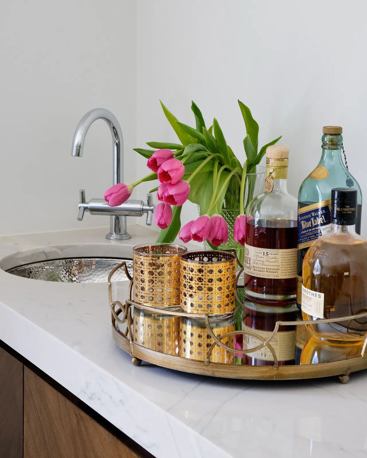 Butler's pantry with marble countertop, sink, wood cabinetry, tray of spirits, gold candle holders, and pink tulips