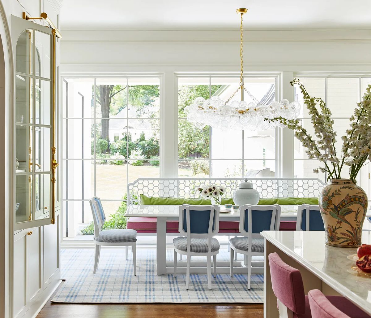 Dining room with white table, upholstered chairs, floral arrangement, and large windows.