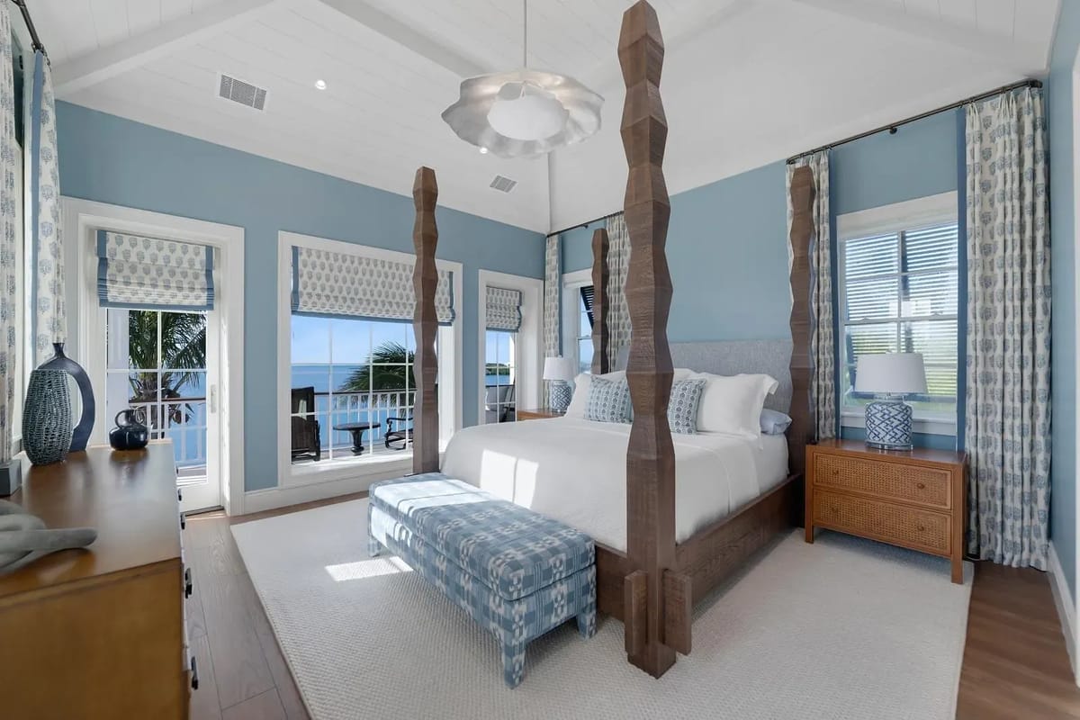 Bedroom with four-poster bed, light blue quilt, nightstands, lamps, large windows, and a dresser