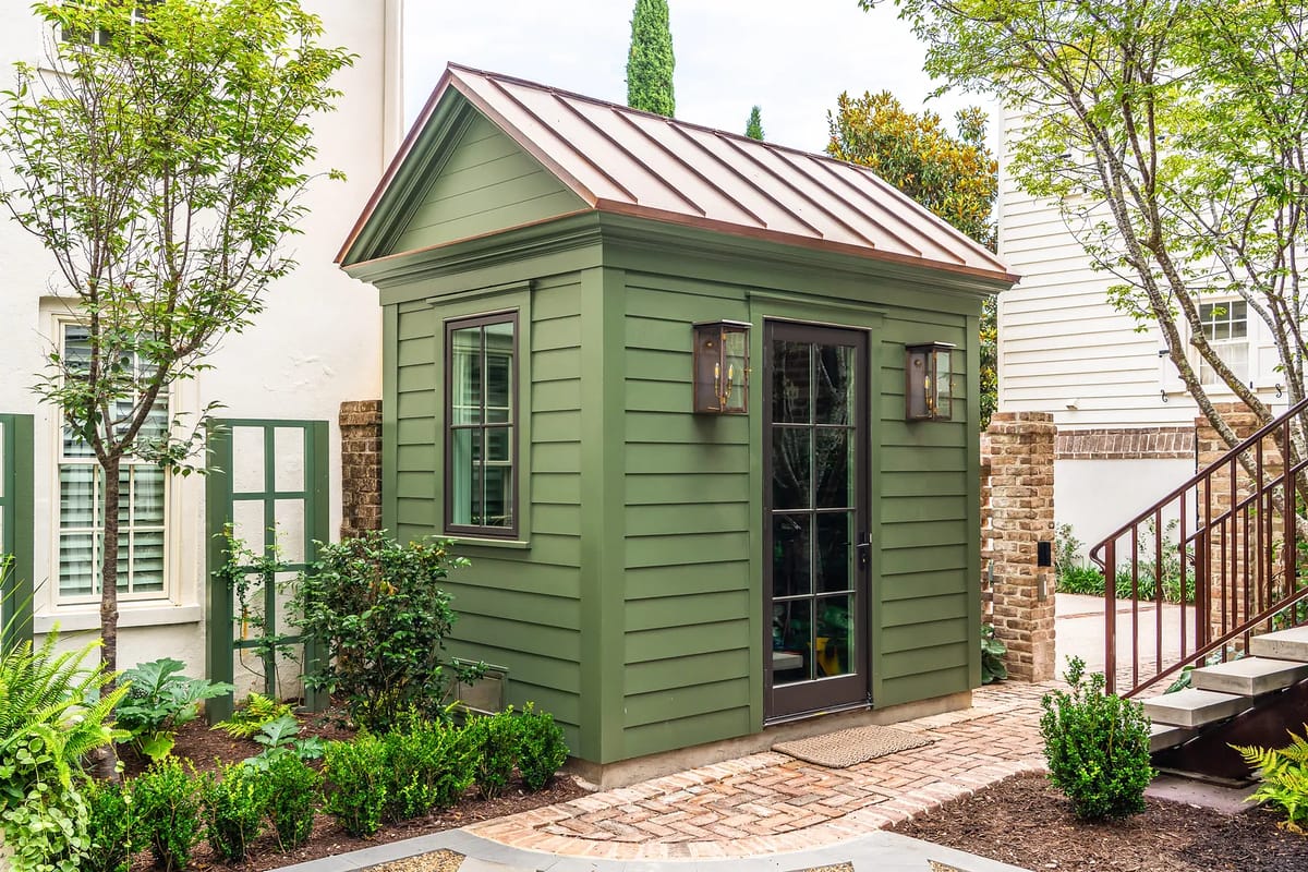 Outdoor shed with green exterior, metal roof, glass door, surrounded by greenery and brick pathways.