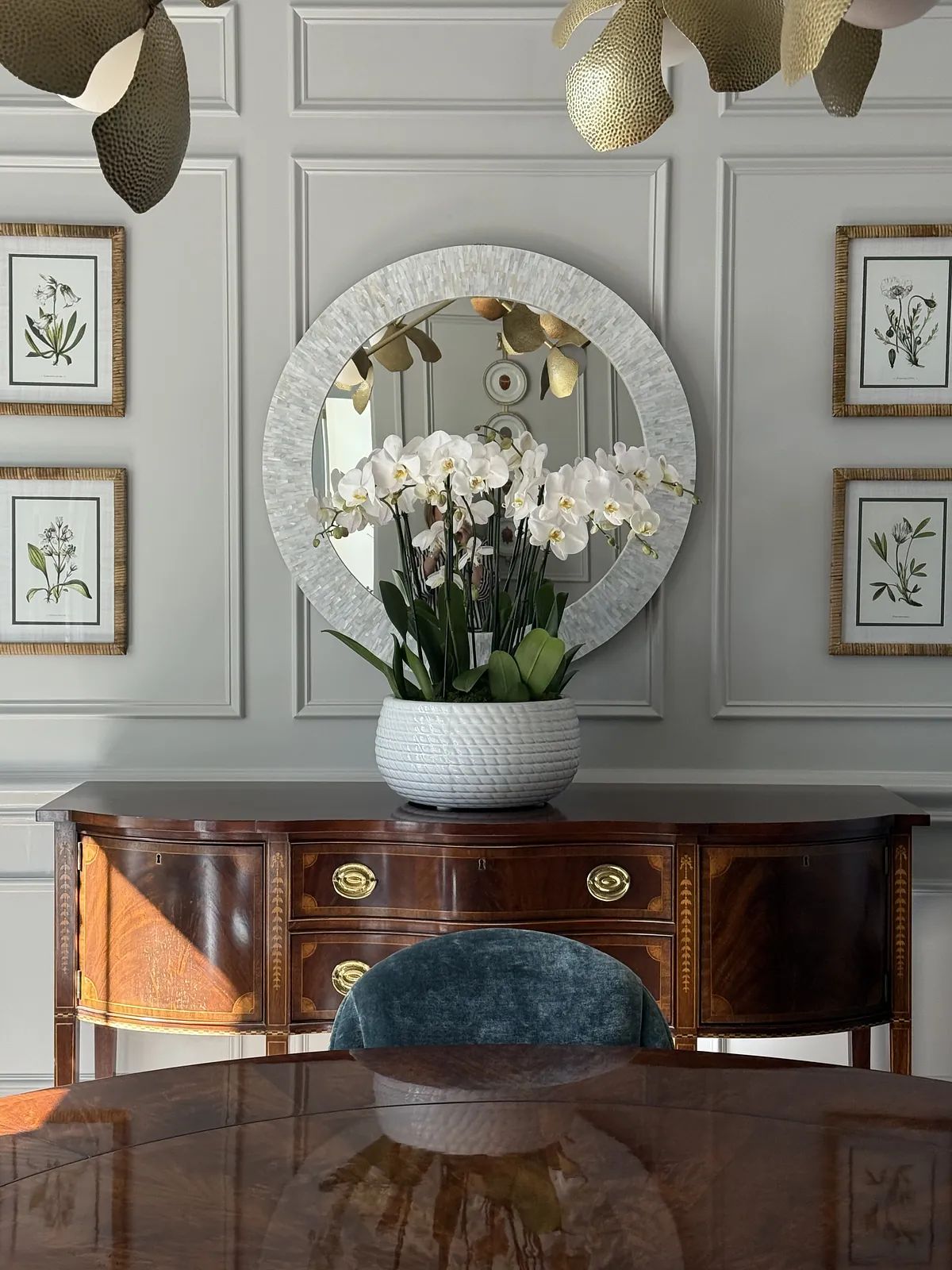 Dining room with round table, upholstered chairs, sideboard, mirror, and botanical prints.