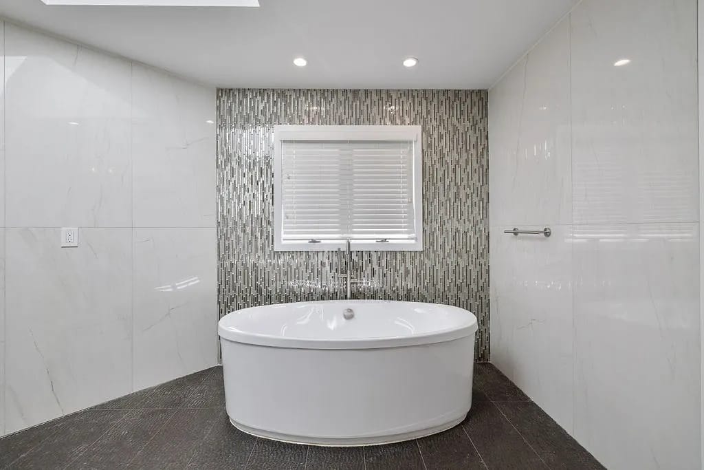 Bathroom with free-standing oval bathtub, wall-mounted faucet, tiled accent wall, and window with blinds