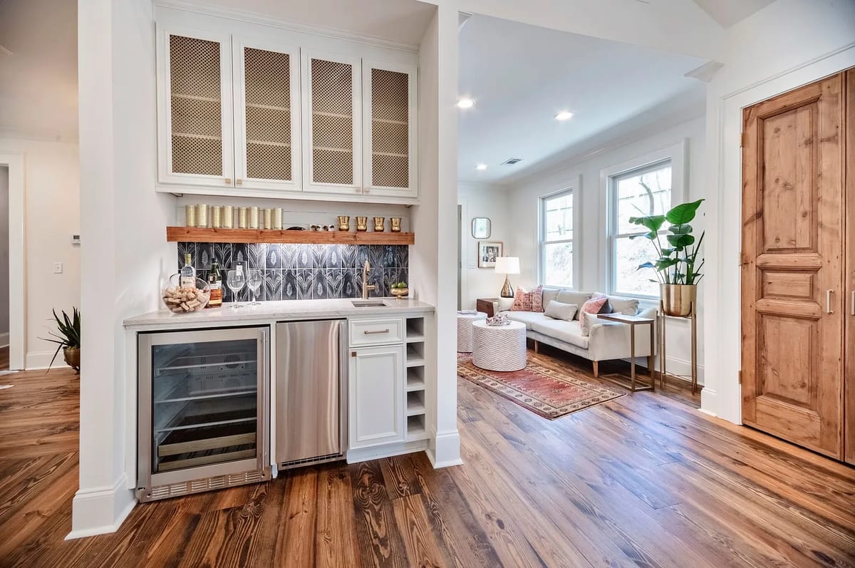 Butler's pantry with wine cooler, wood cabinetry, backsplash, and adjacent living area with sofa and rug.
