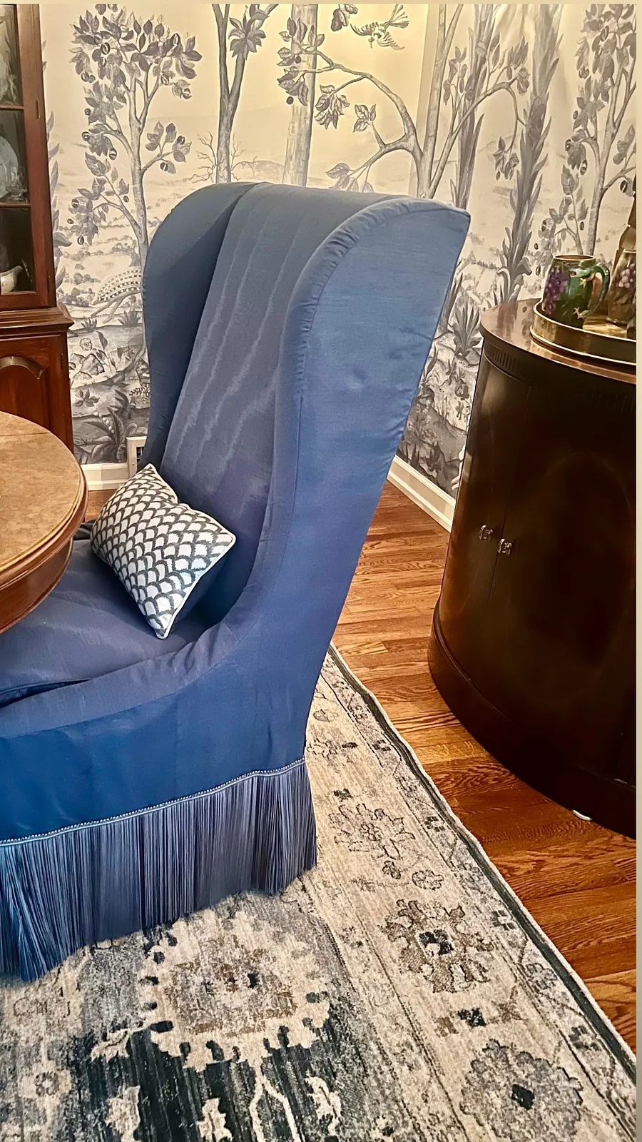 Dining room with round table, blue chair, accent pillow, floral wallpaper, and dark wood cabinet.