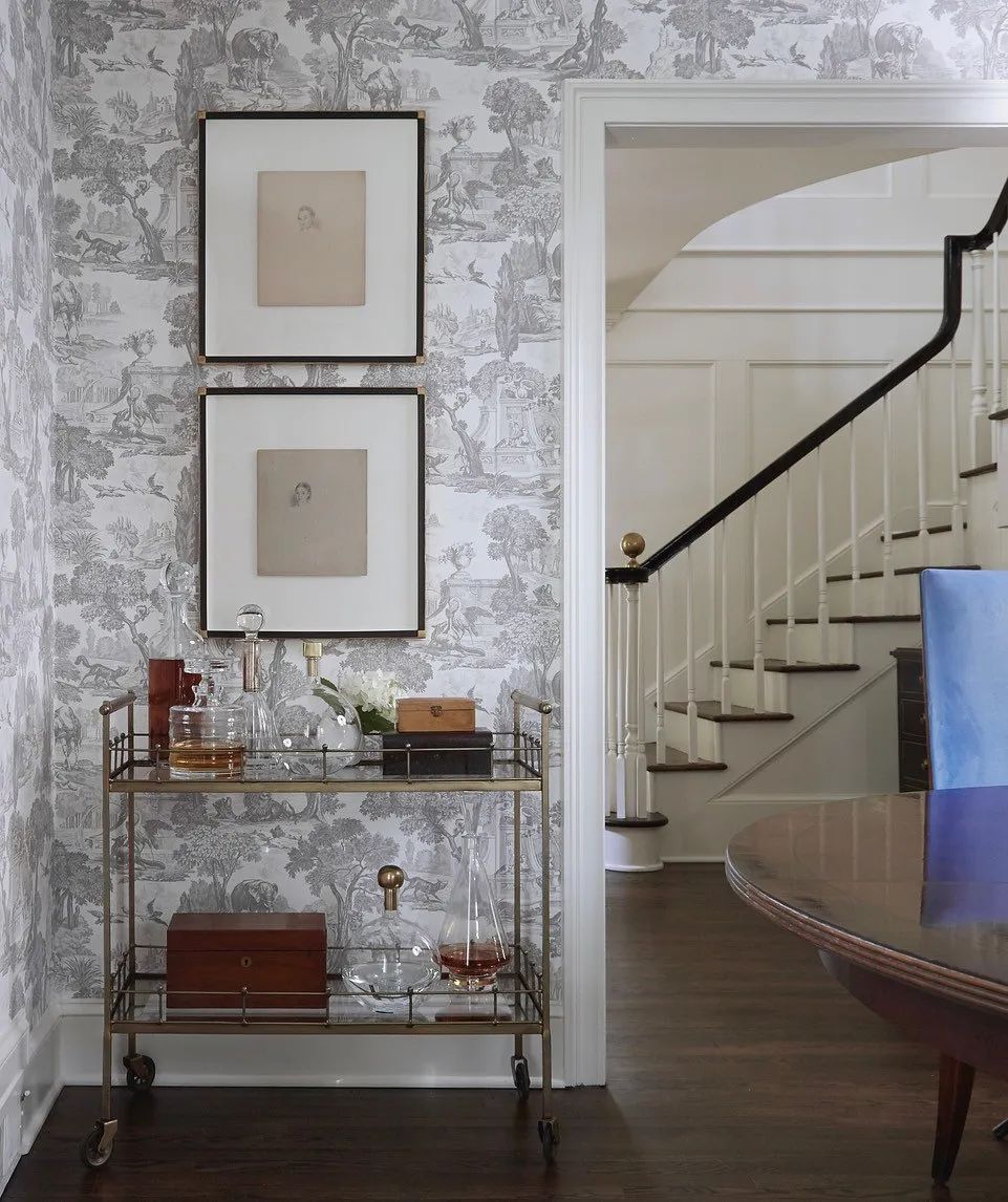 Foyer with patterned wallpaper, glass bar cart, artwork, staircase, dining table, and blue chair