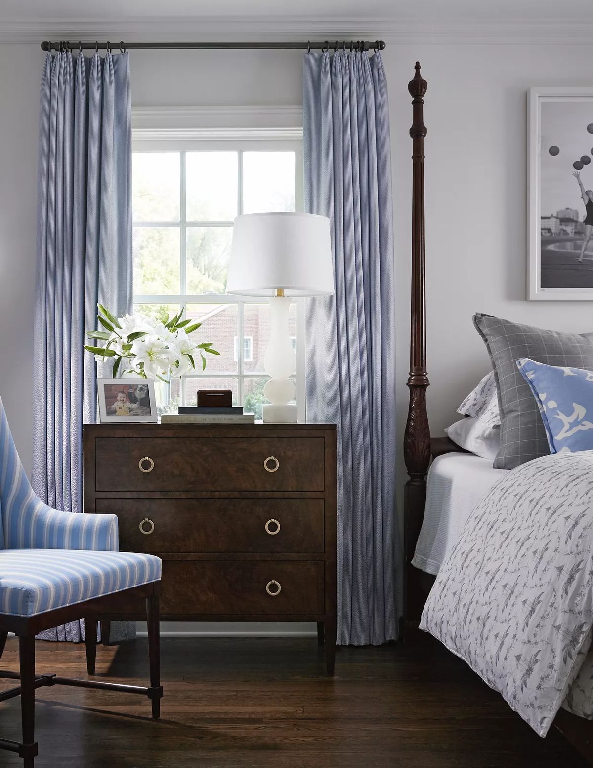 Bedroom with four-poster bed, bedside table, blue striped chair, and white flower vase by the window