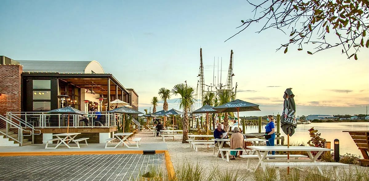 Exterior area with picnic tables, umbrellas, palm trees, and a brick building by a water body.
