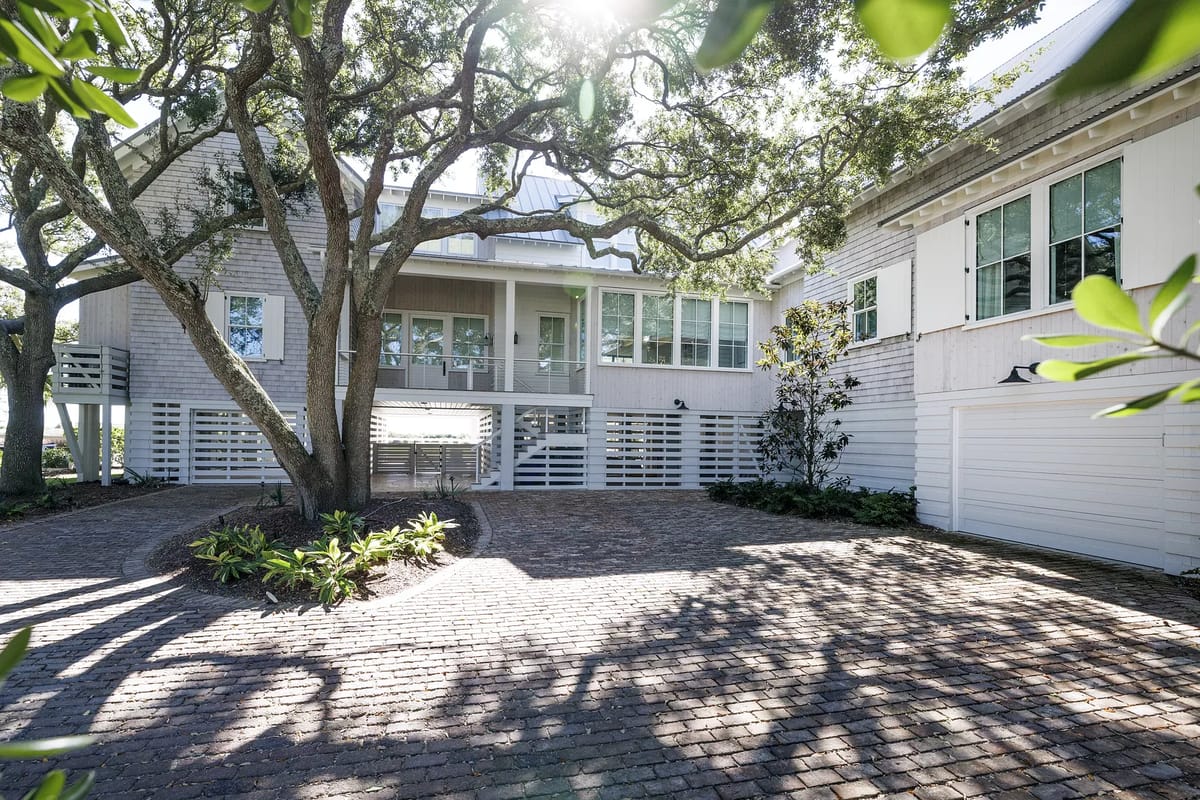 Front exterior of a house with brick driveway, shrubs, large windows, elevated porch, and mature trees.