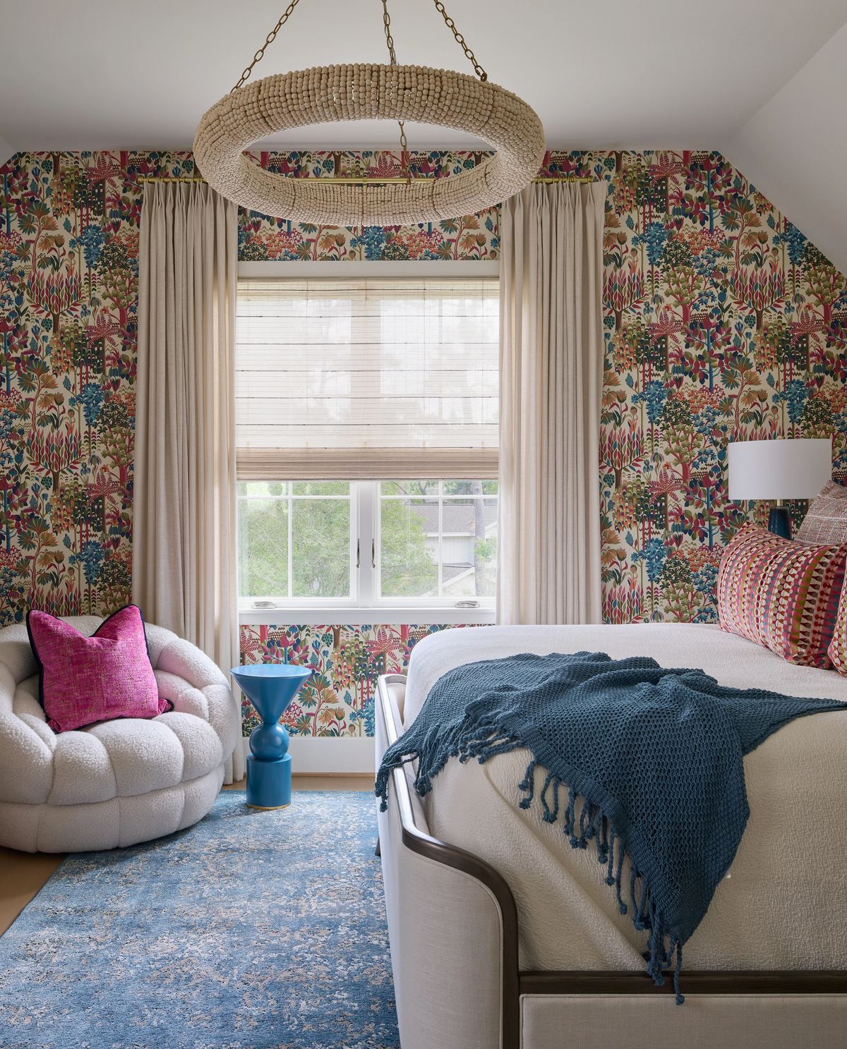 Bedroom with large bed, floral wallpaper, upholstered chair, blue side table, and large window.