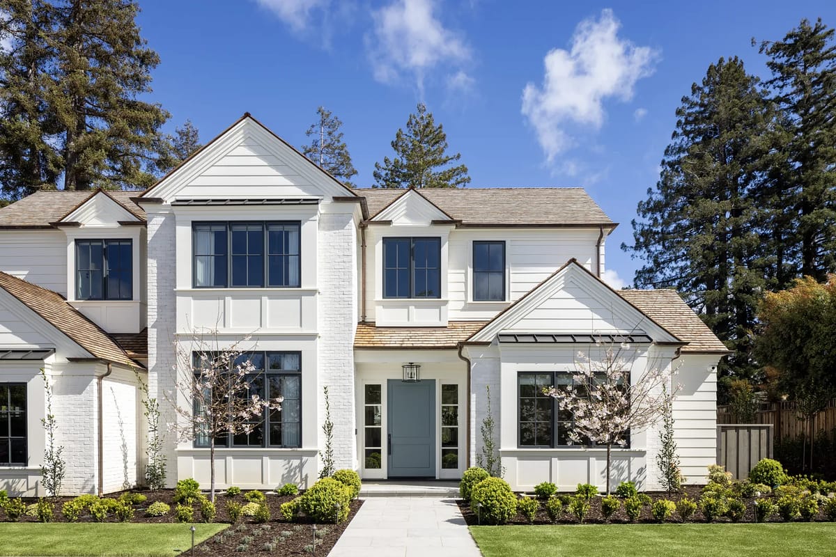 Front exterior of a house with white brick, large windows, glass door, and landscaped entrance
