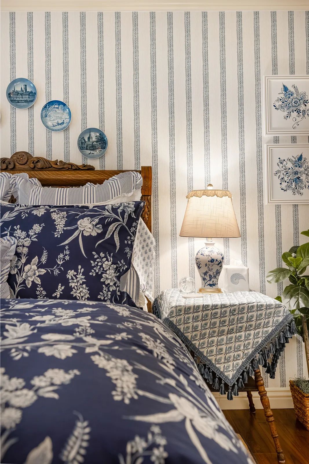 Bedroom with wooden bed, navy bedding, nightstand with lamp, striped wallpaper, and blue decorative plates.