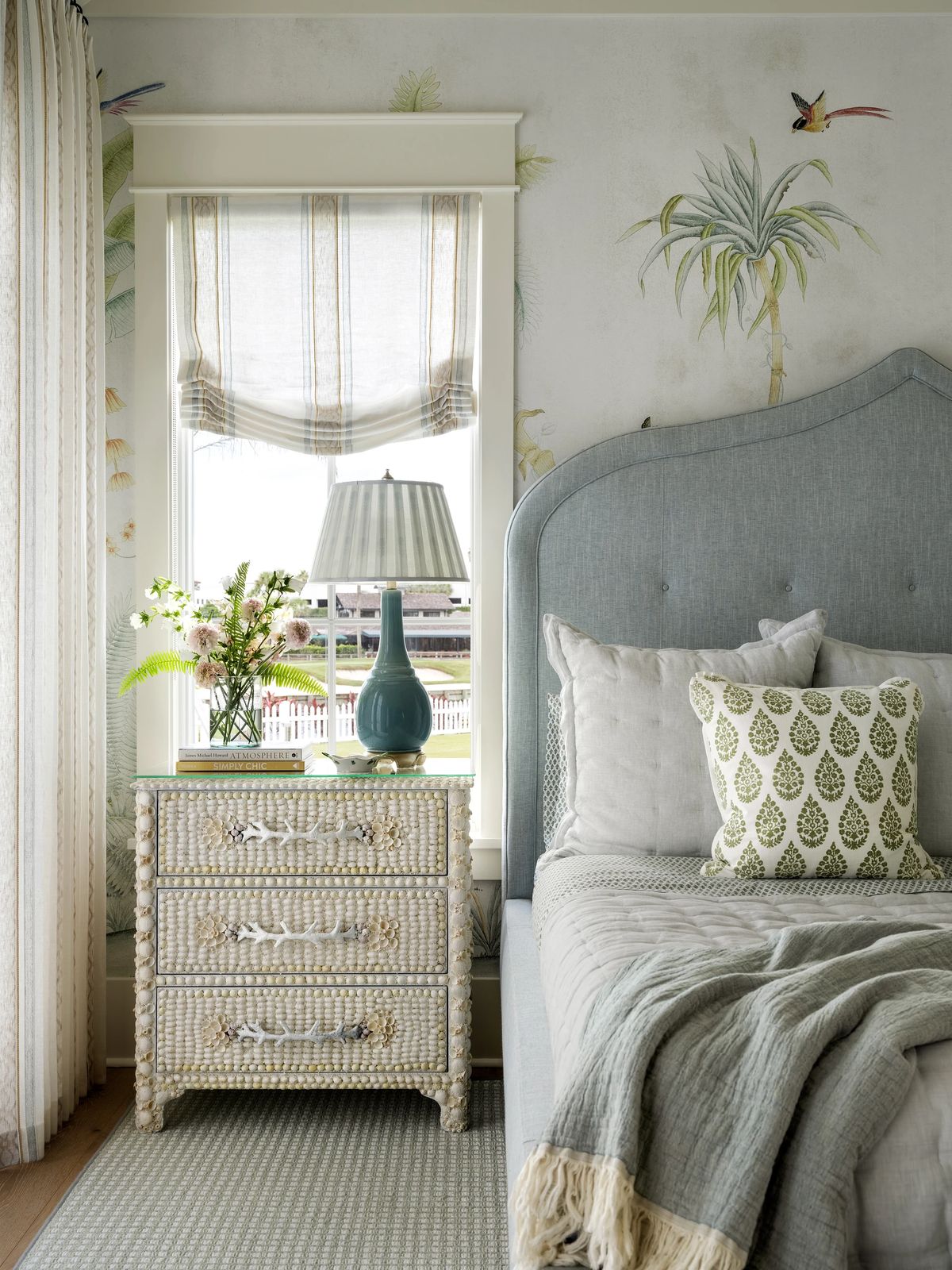 Bedroom with blue headboard, quilt, woven chest, lamp, botanical wallpaper, and window with curtains