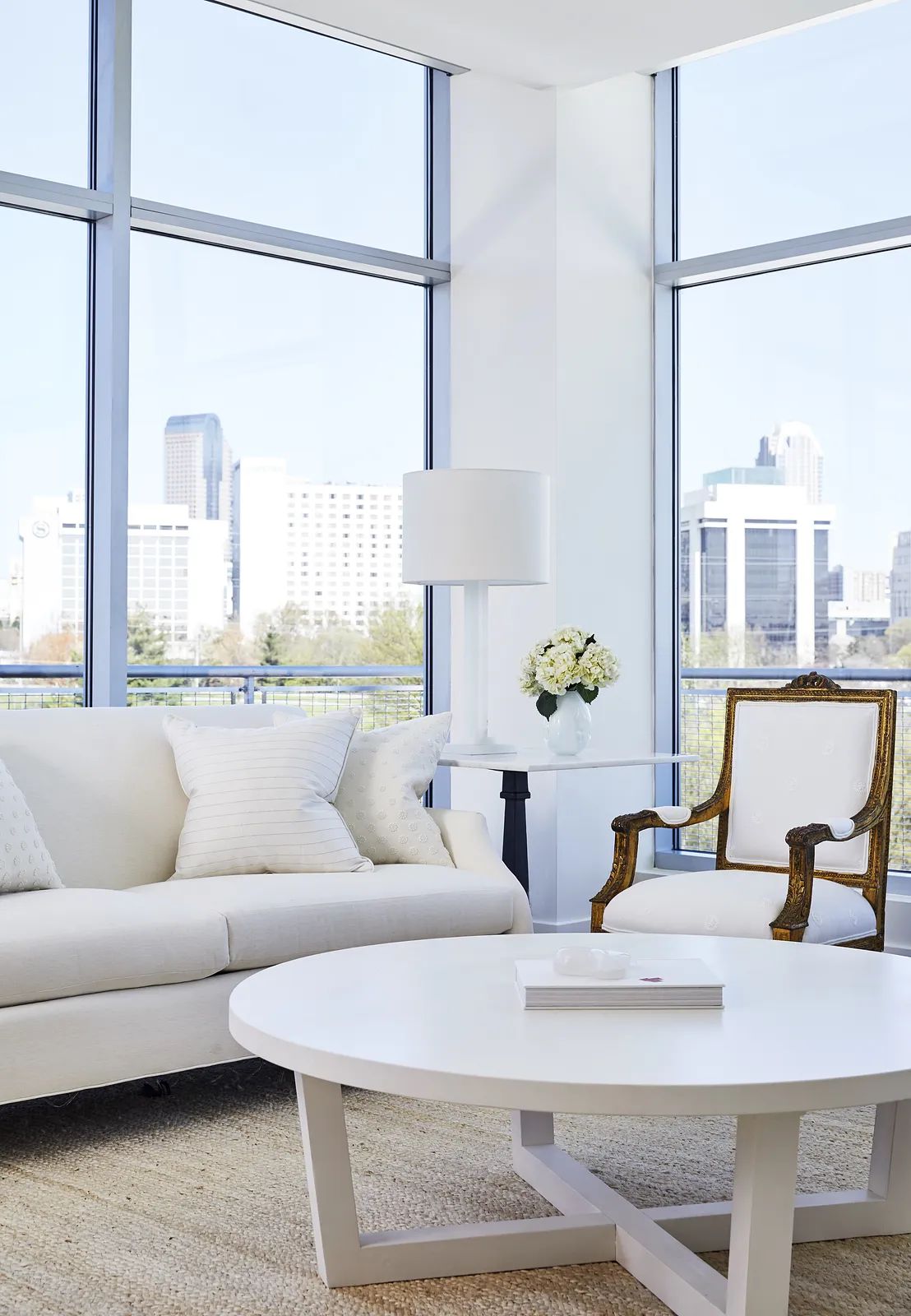 Living room with white sofa, round coffee table, gold-accented chair, floor lamp, and large glass windows.