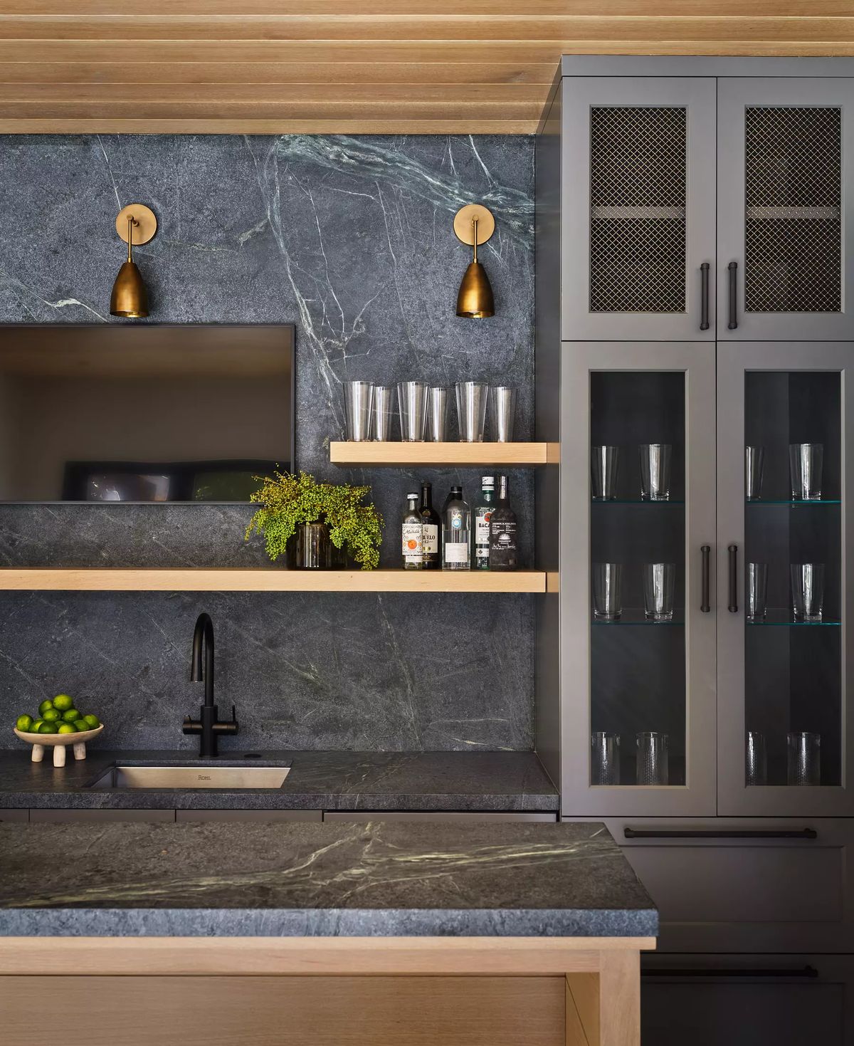 Kitchen with dark stone countertop, gray cabinets, open shelving, sink, and glass-front cabinets with wall sconces