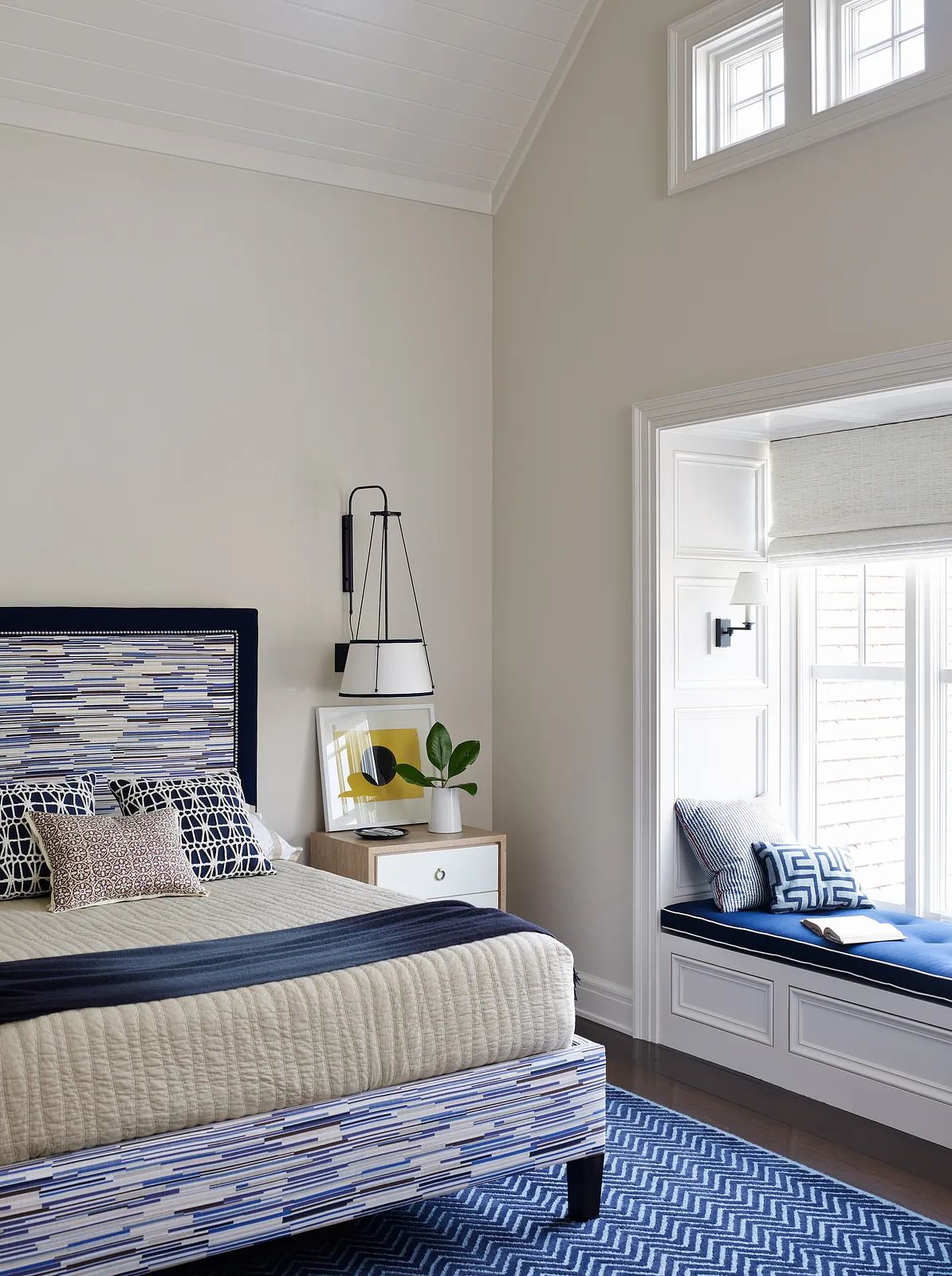 Bedroom with patterned headboard, quilted bedding, nightstand, window seat, and blue area rug.