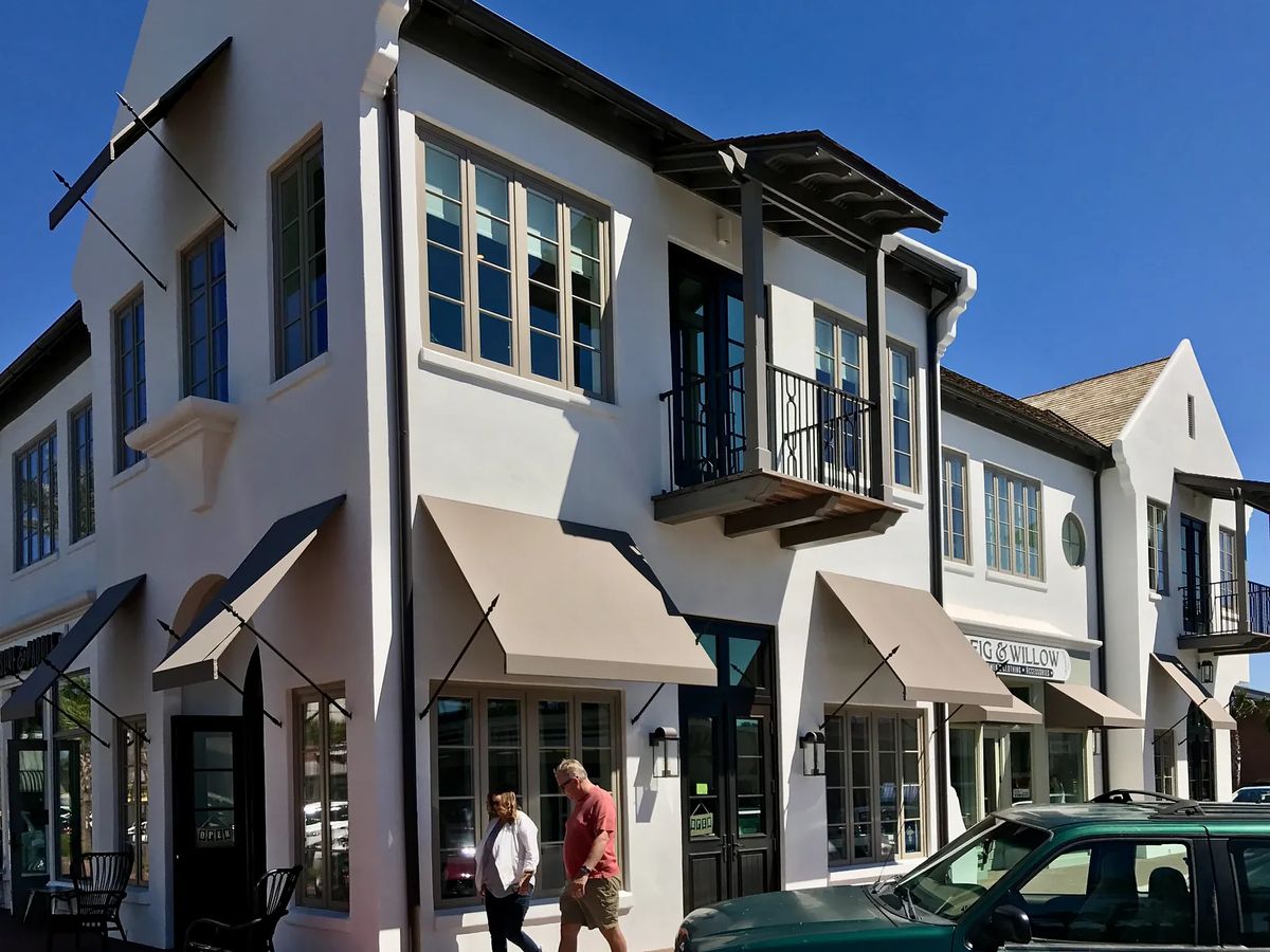 Two-story building with white stucco, large windows, balcony, awnings, pedestrians, and parked car.