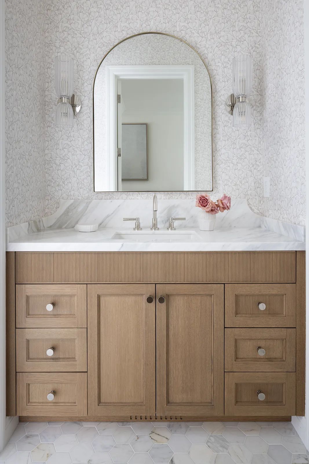 Bathroom with wooden vanity, marble countertop, round mirror, floral wallpaper, and wall sconces.