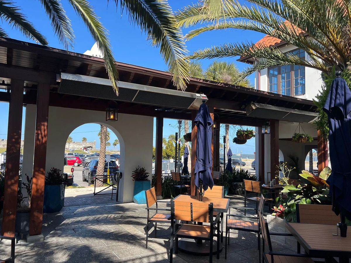 Outdoor patio with wooden tables, chairs, umbrellas, potted plants, and a view of water.