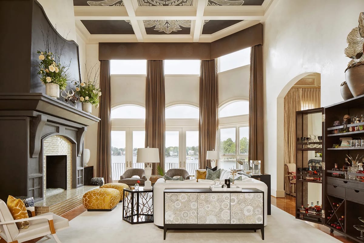 Living room with large fireplace, beige sofa, patterned ottoman, glass coffee table, and tall windows overlooking water.