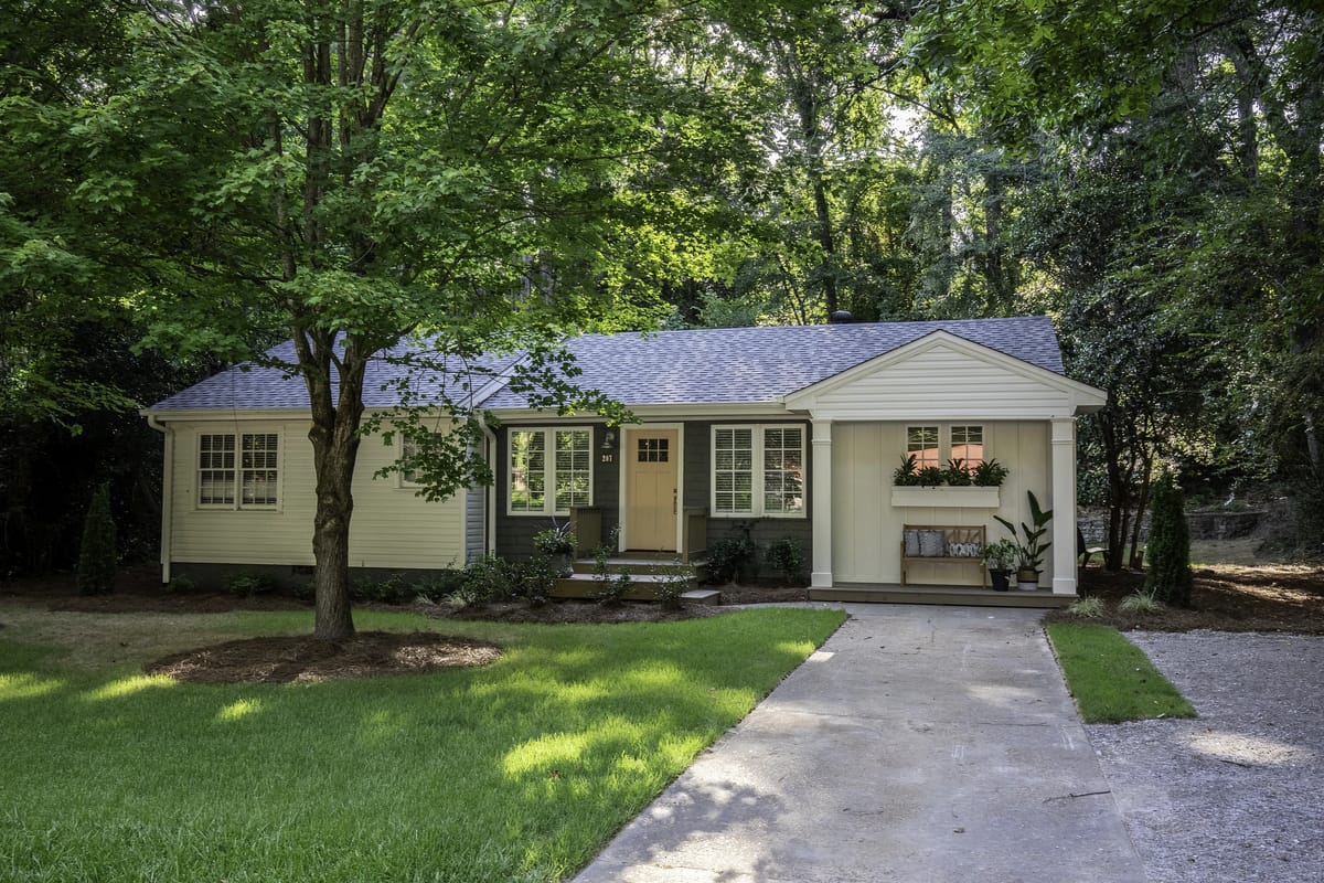 Front exterior of a single-story house with light yellow siding, gray roof, concrete driveway, and landscaping.