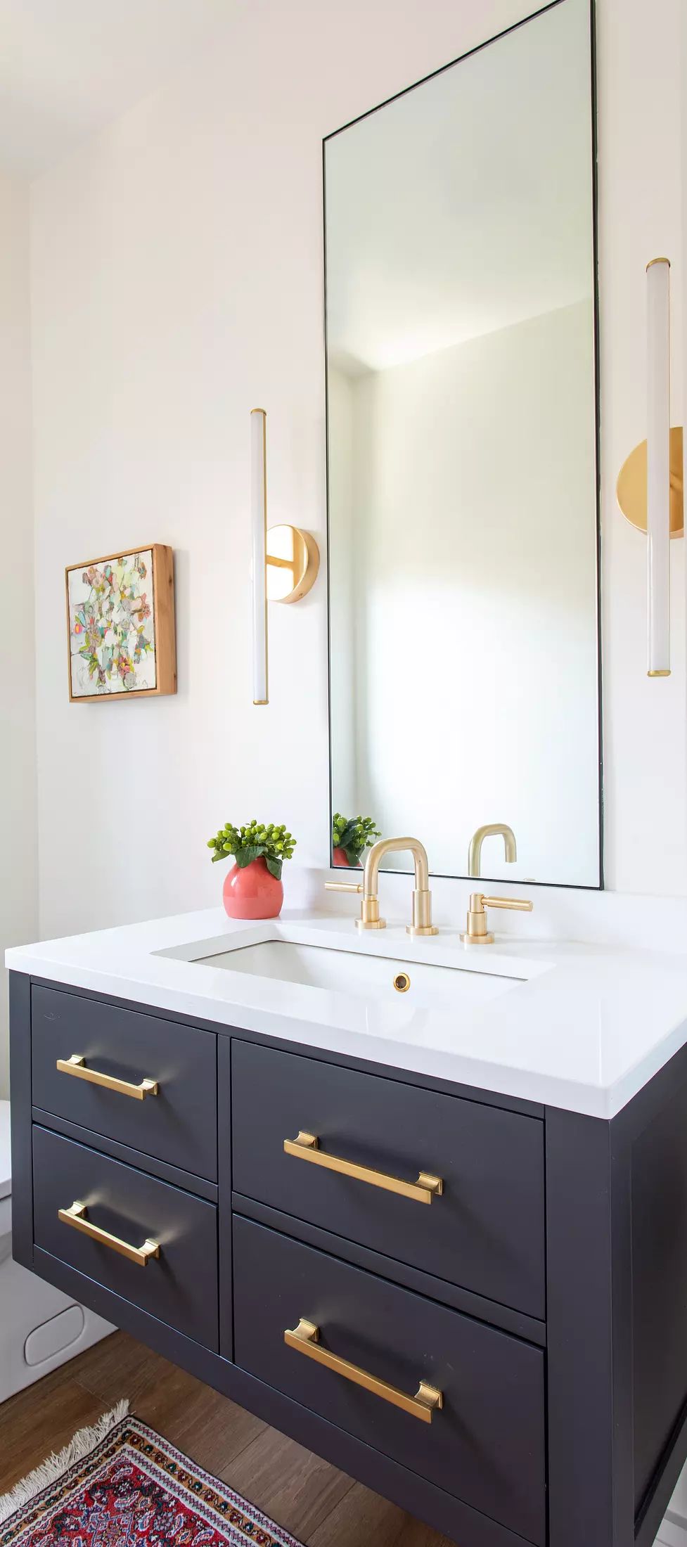 Bathroom with dark cabinet, white countertop, large mirror, sconces, small plant, and colorful rug