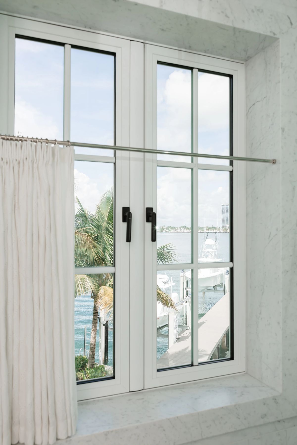 Bathroom with large window, white curtain, and light-colored stone walls overlooking palm trees and water.