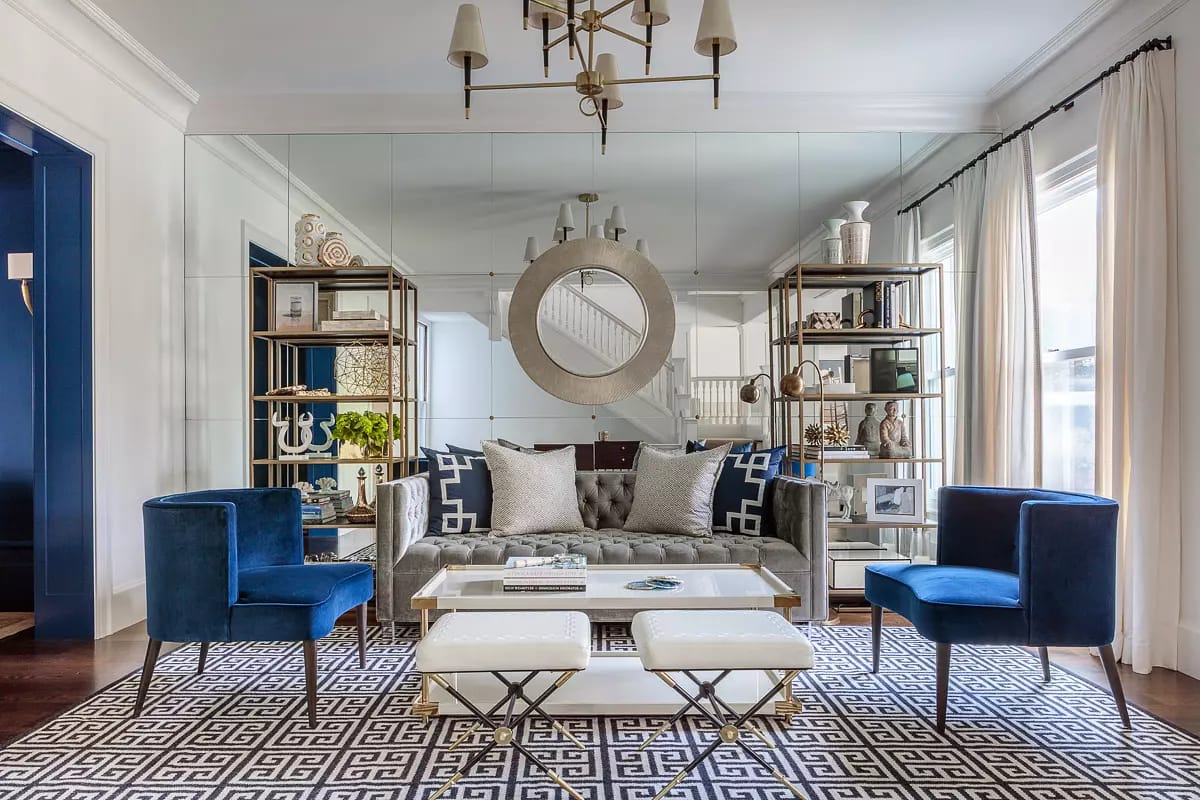 Living room with gray sofa, blue chairs, glass coffee table, shelves, round mirror, and large windows.