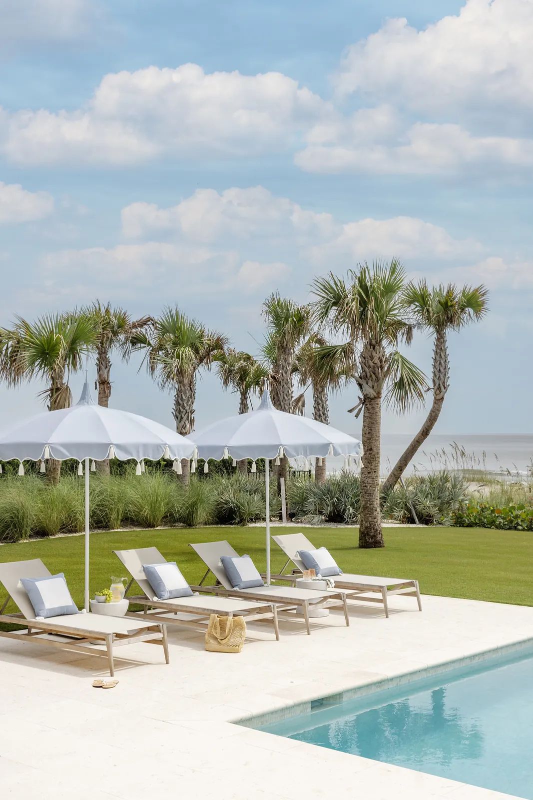 Patio area with lounge chairs, umbrellas, swimming pool, grass, and palm trees by the beach.