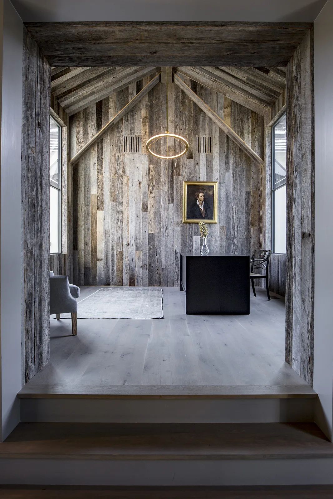 Office with wooden wall, black desk, chair, painting, exposed beams, and light-colored wood flooring.