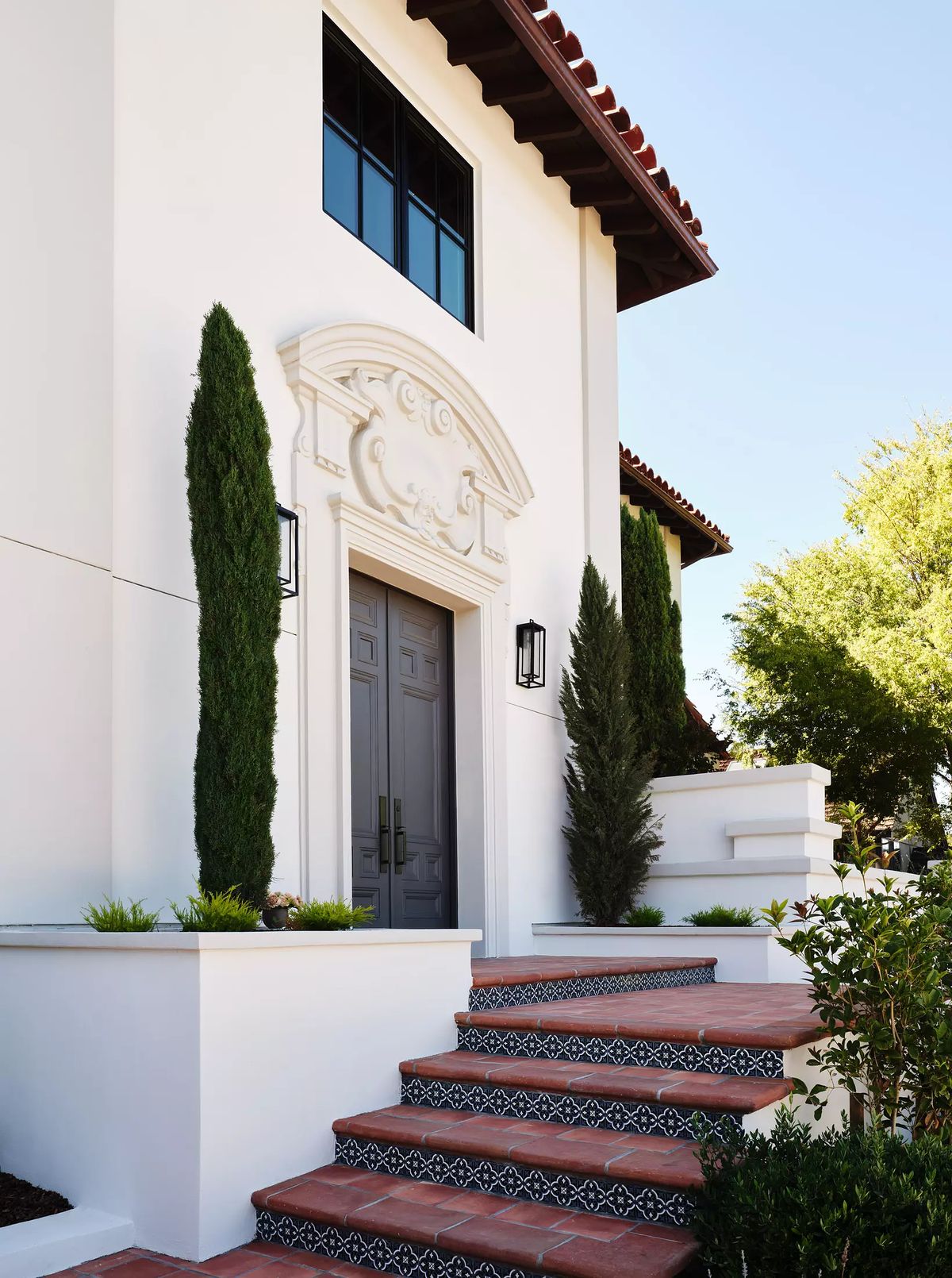 Exterior front view with decorative entrance, terracotta steps, cypress trees, and low shrubs.