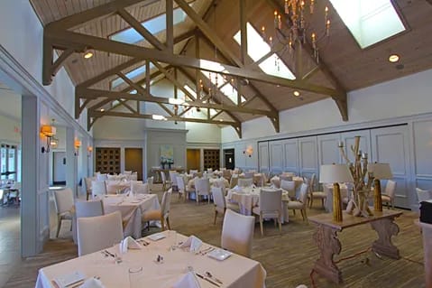 Dining room with round tables, white tablecloths, wooden beams, skylights, and upholstered chairs.