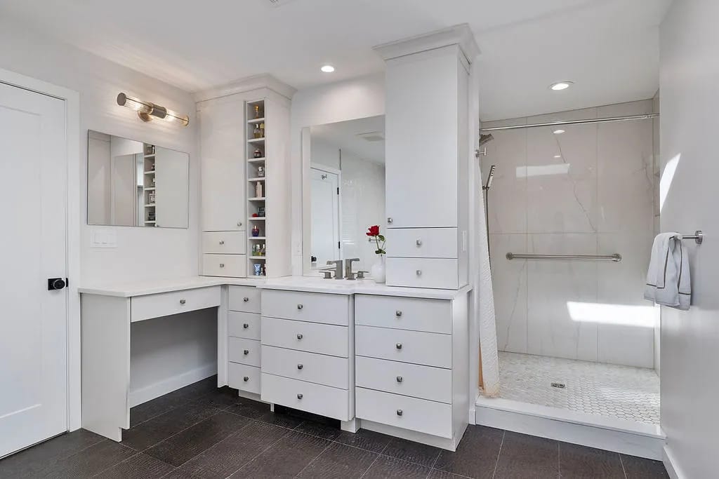 Bathroom with white vanity, mirror, built-in shelves, walk-in shower, and dark tile flooring with natural light.