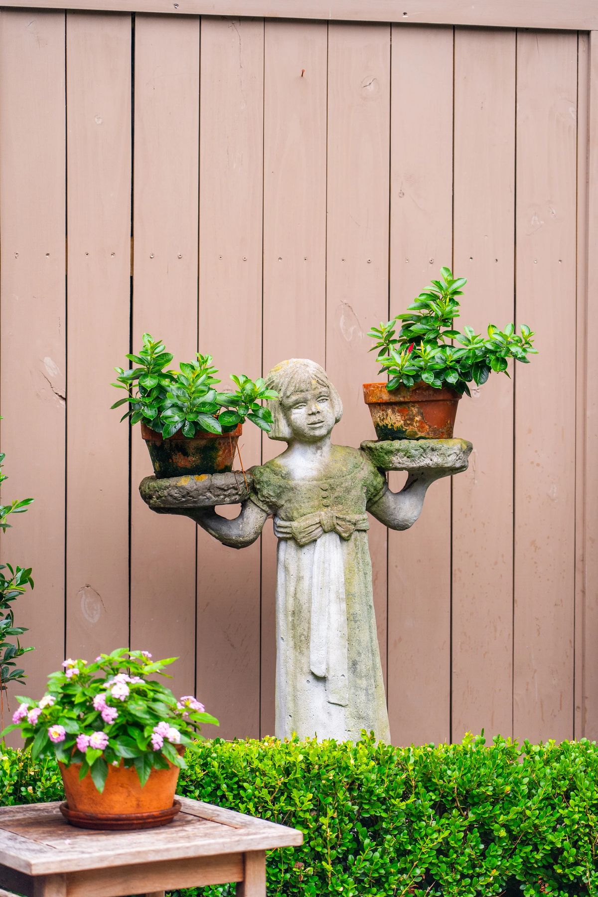 Garden with stone statue holding potted plants, wooden table, and flowering plants against a wooden fence.