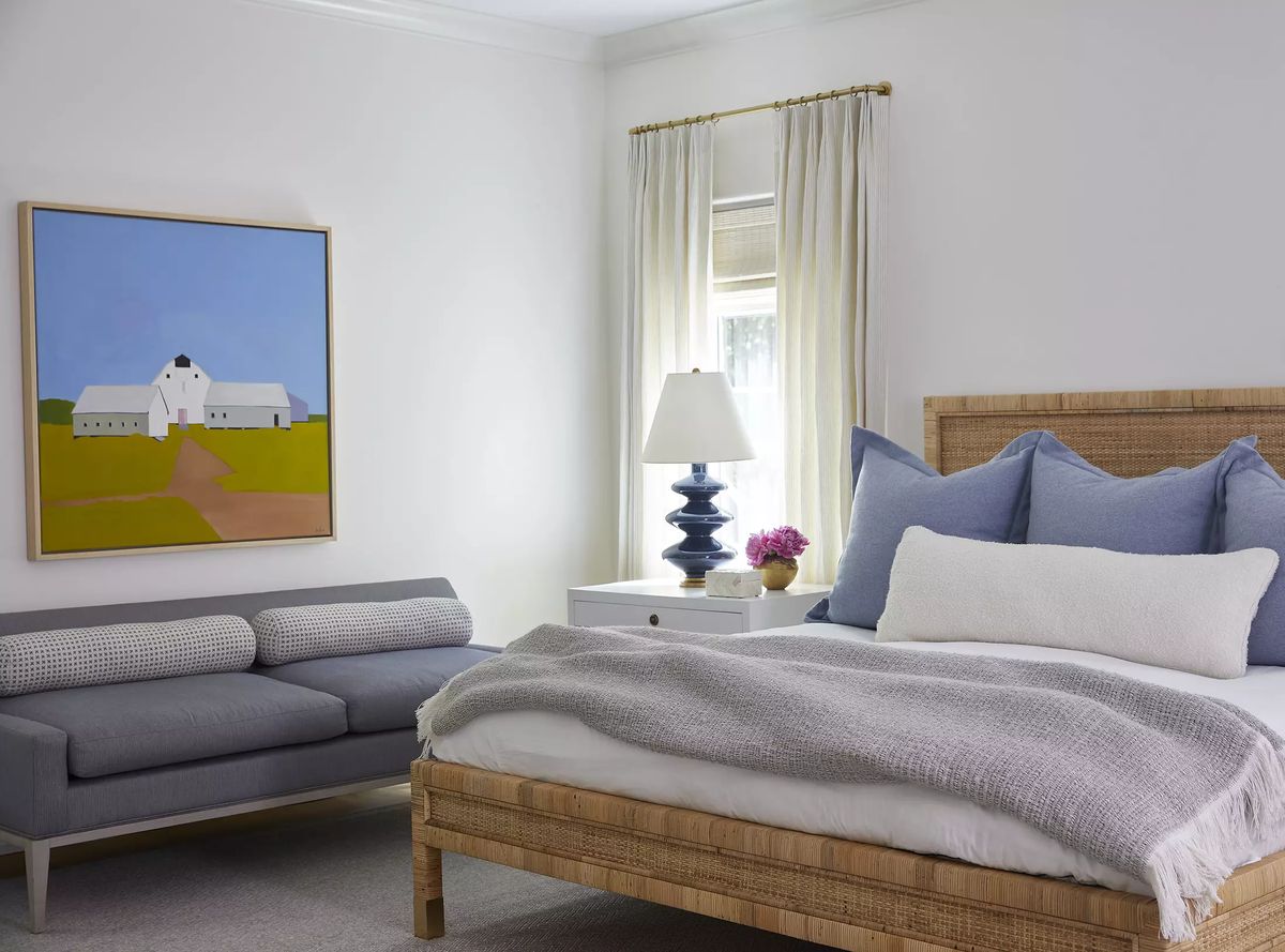 Bedroom with woven headboard bed, gray sofa, accent pillows, lamp, and large framed painting.
