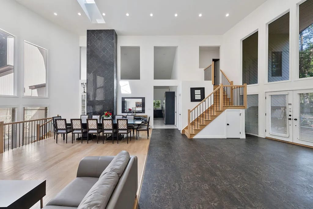 Living room featuring gray sofa, dining area, large windows, stair, and stone fireplace column with natural light