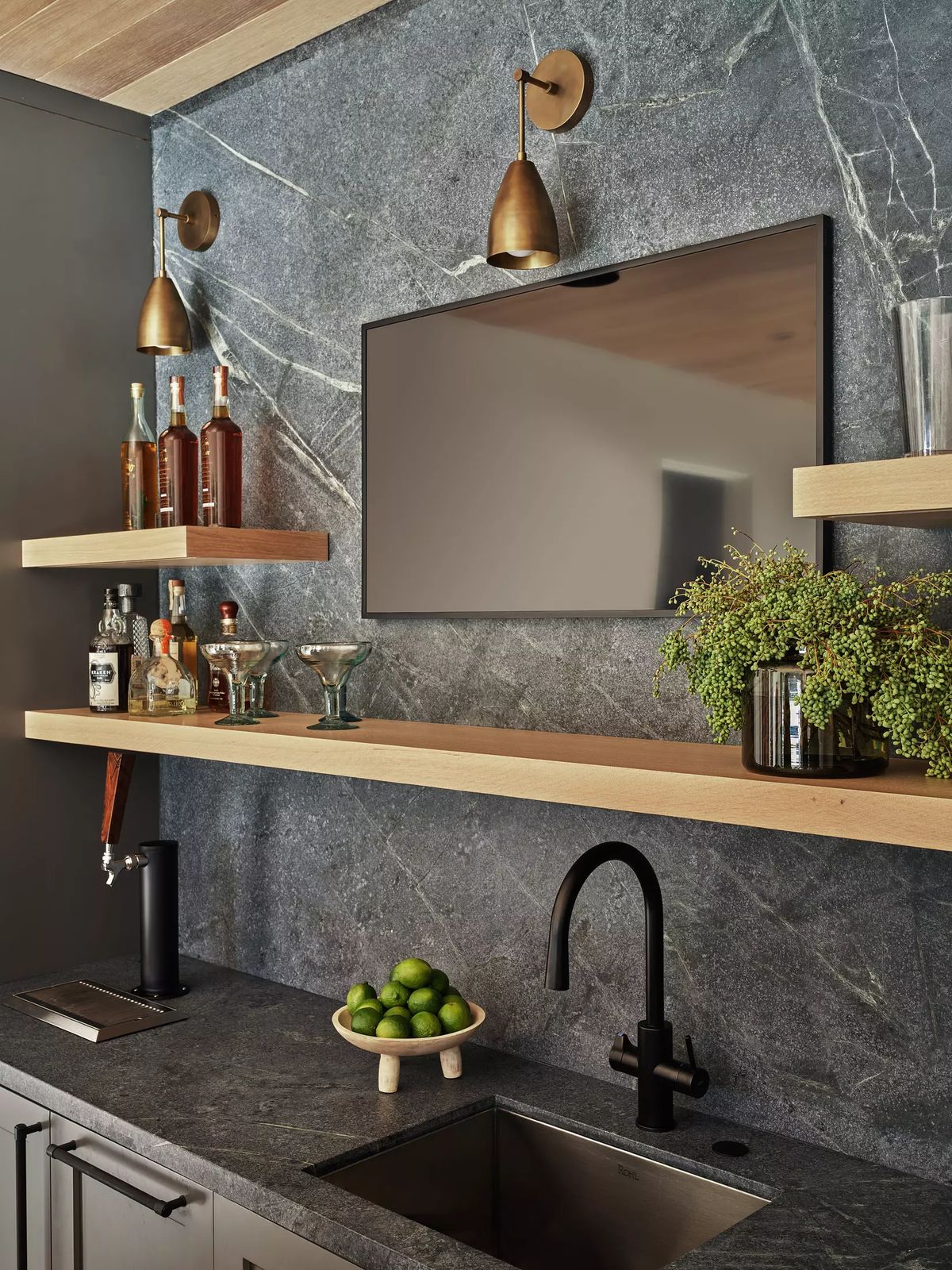 Kitchen area with black countertop, mounted shelves, sink, wall-mounted TV, and limes in a bowl.