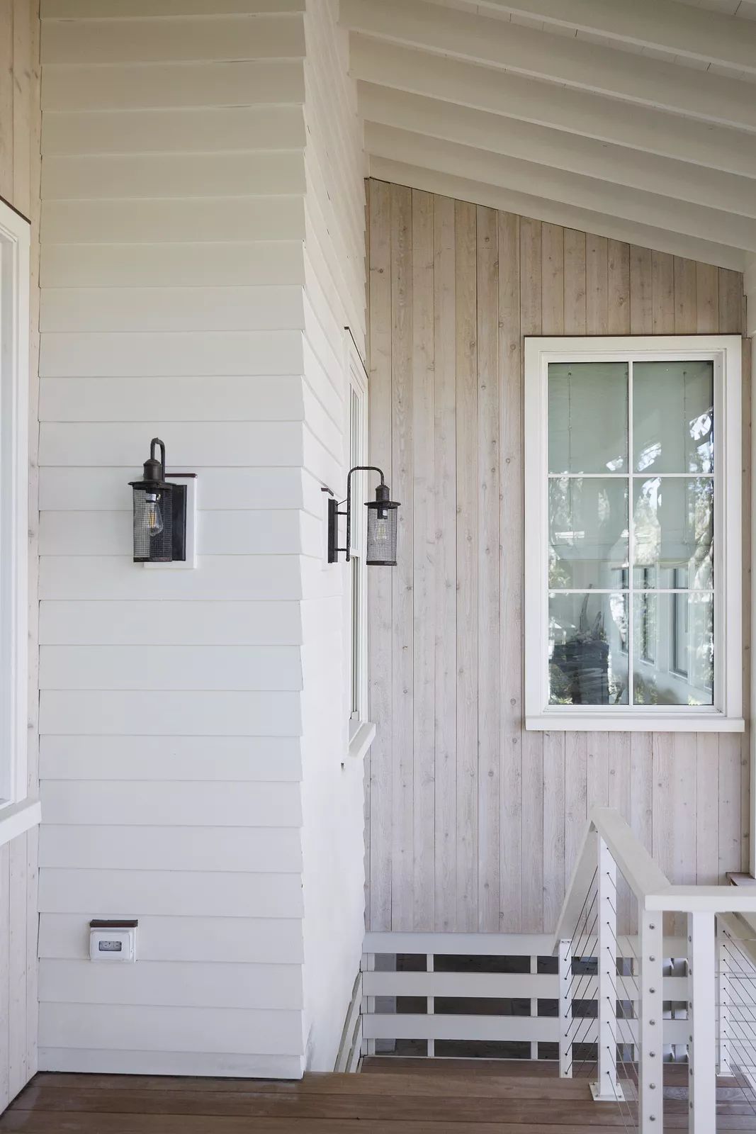 Exterior area with white siding, wood paneling, two wall sconces, and large windows.