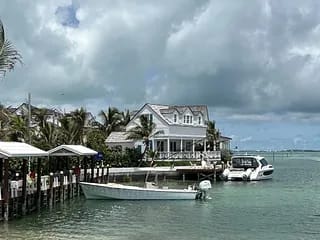 Waterfront property with two-story white house, boats, palm trees, and outdoor seating area on the dock.