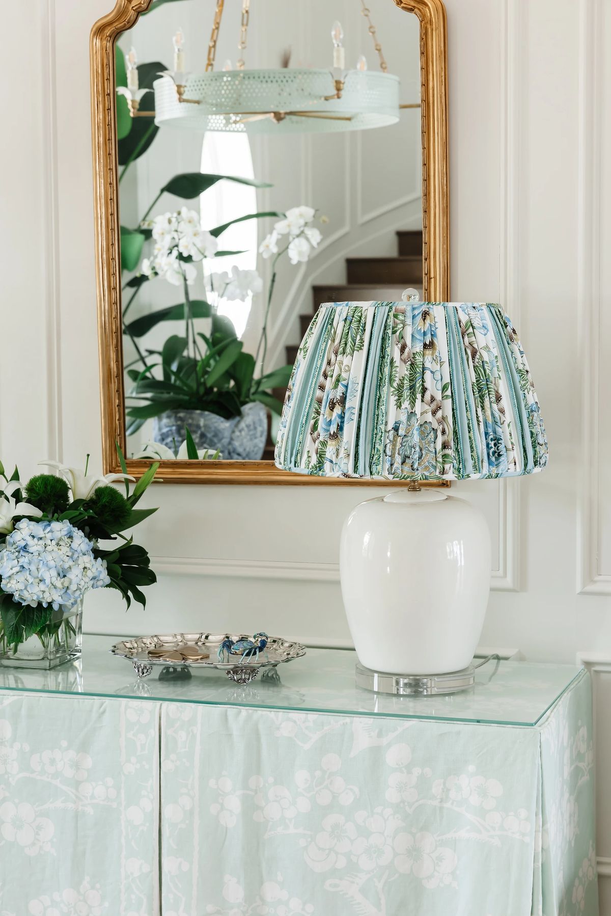 Foyer with glass console table, floral lamp, vase of hydrangeas, and large round mirror with staircase in background