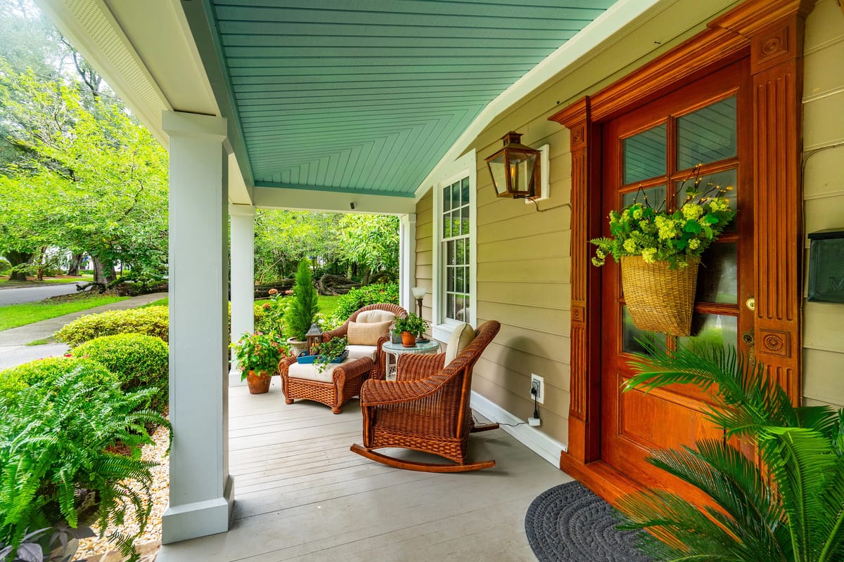 Front porch with wicker chairs, small table, potted plants, large wooden door, and blue ceiling.
