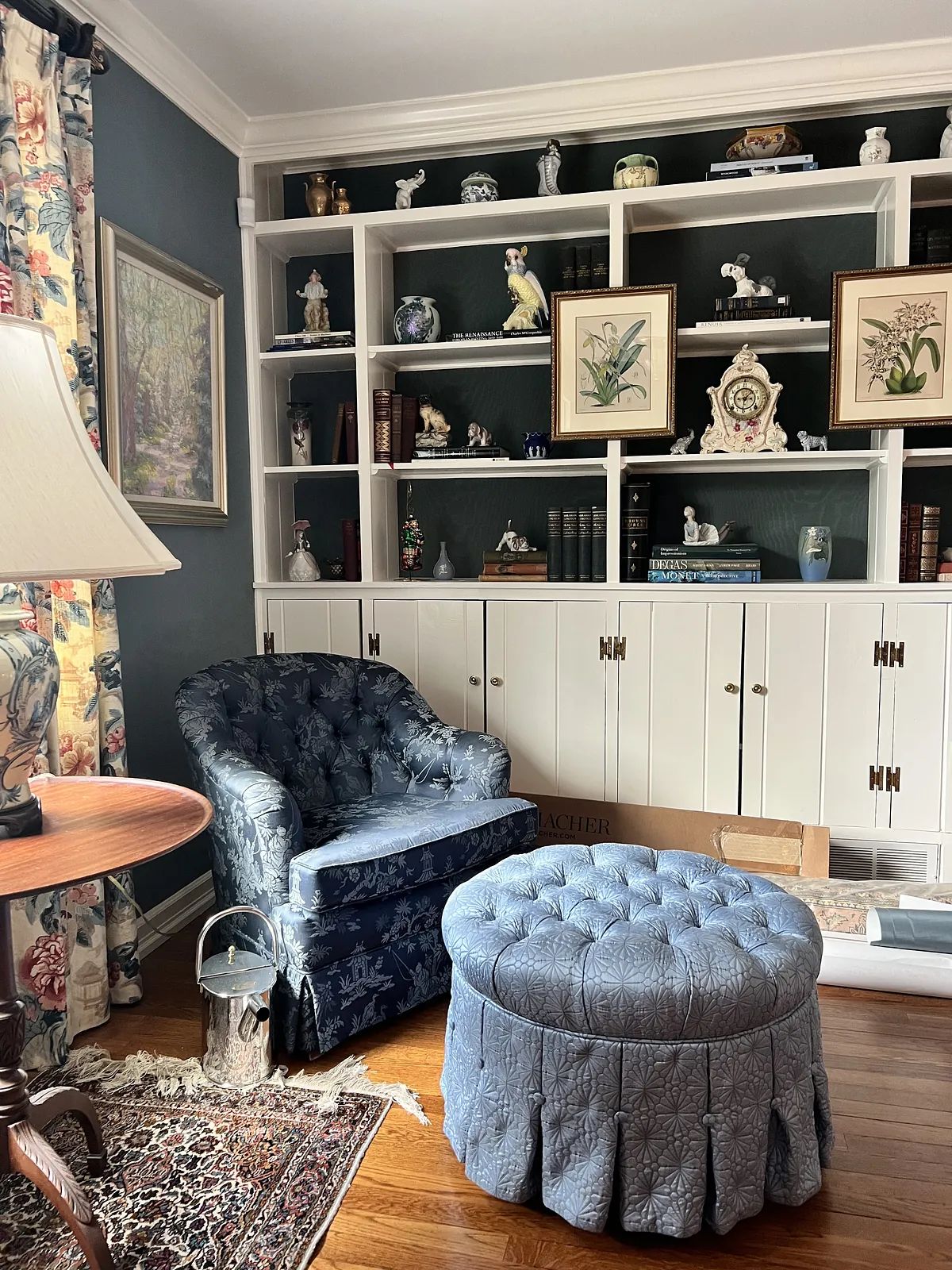 Living room with blue armchair, round ottoman, wooden side table, bookcase, and floral curtain.