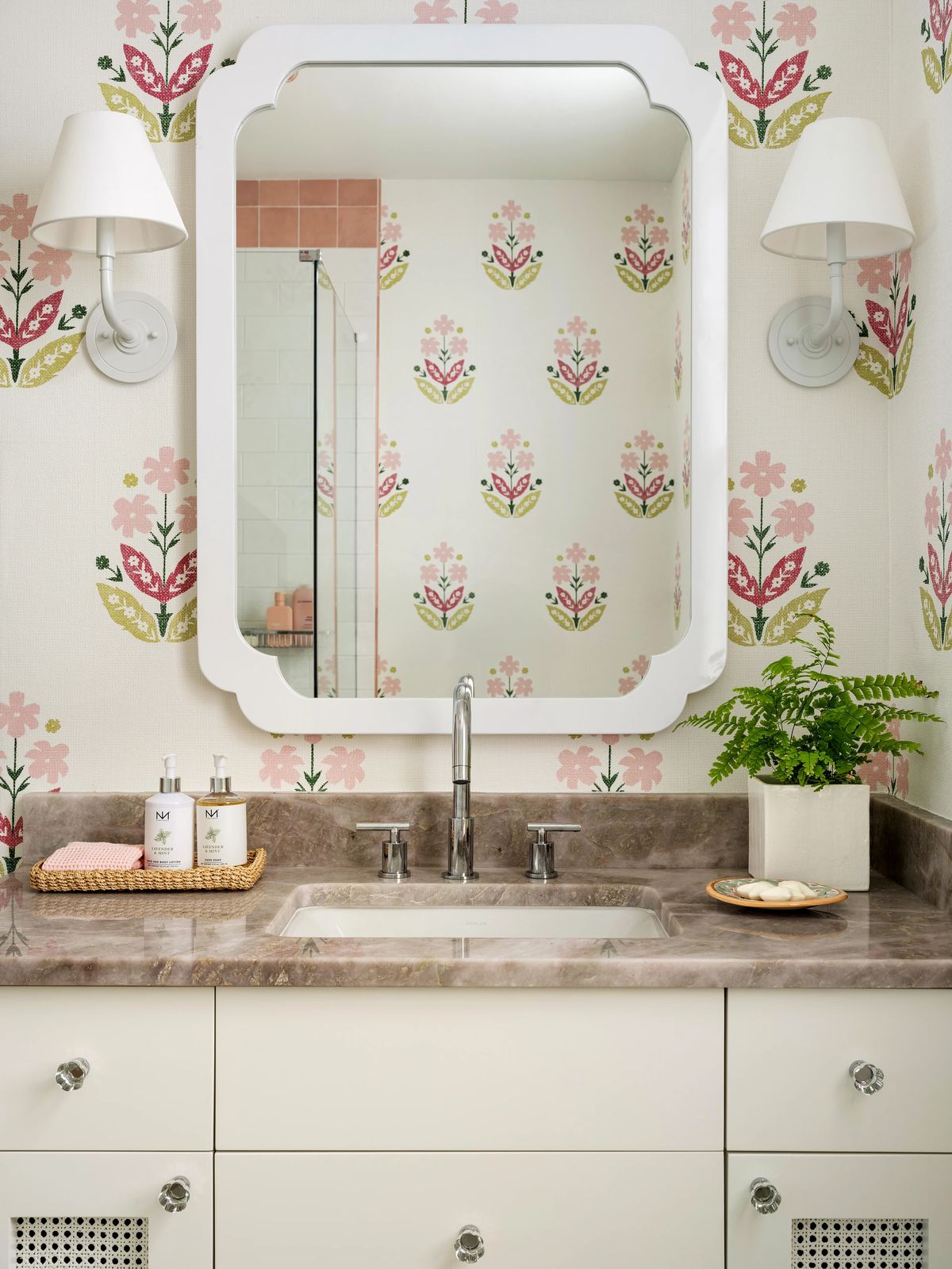 Bathroom with a sink, mirror, floral wallpaper, decorative bottles, and a potted plant.